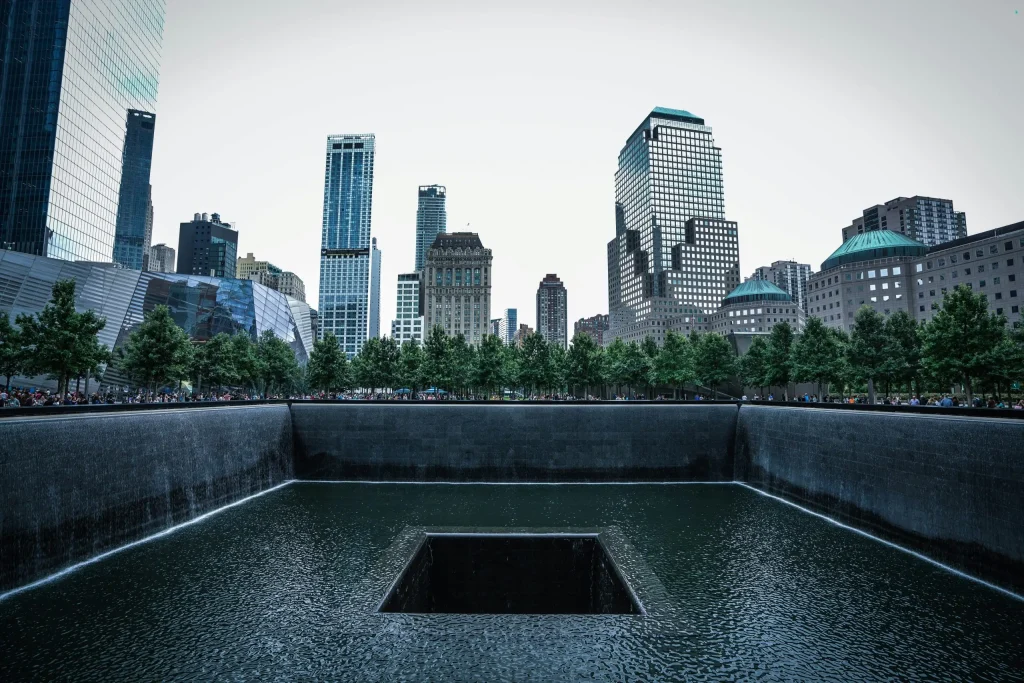 A view of the 9/11 Memorial in New York City, featuring the North Pool surrounded by trees and framed by the city's skyscrapers. The reflective pool, with its cascading water, serves as a somber tribute to the events of September 11, 2001.