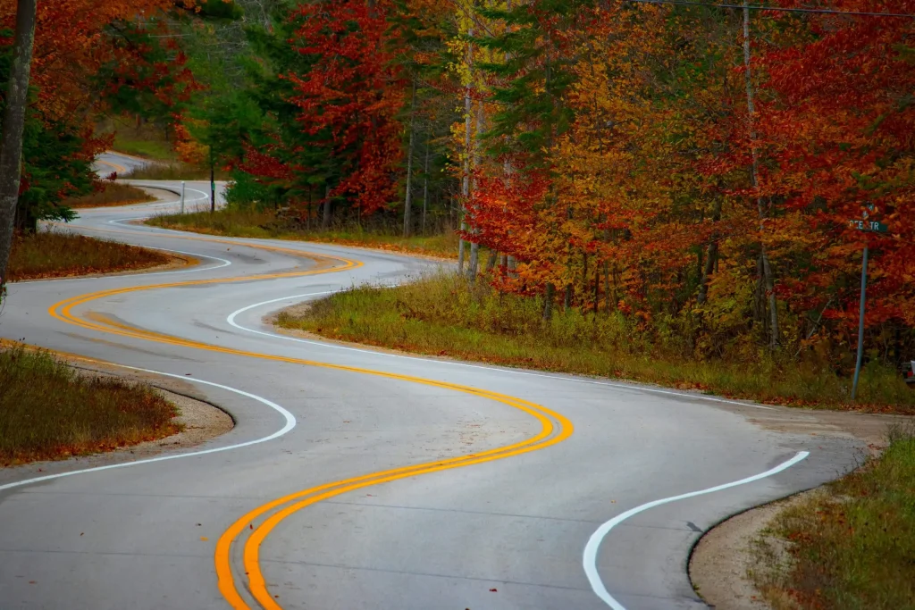 A winding road through Door County, Wisconsin, flanked by vibrant autumn foliage. The trees on either side of the road showcase a beautiful array of red, orange, and yellow leaves, making it a picturesque drive during the fall season.