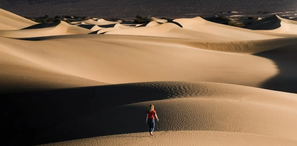 A lone traveler walks barefoot across vast, golden sand dunes under a warm, setting sun. The expansive desert landscape creates a dramatic and serene atmosphere, with shadows accentuating the undulating dunes.
