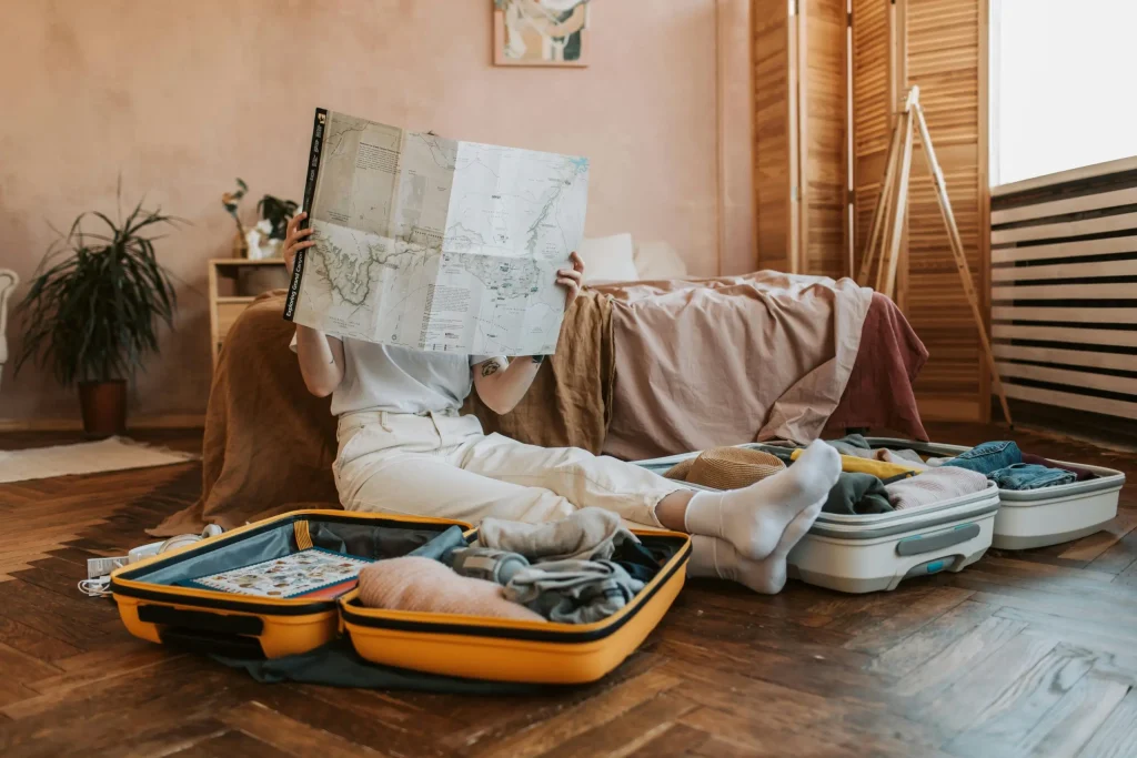 A person sitting on the floor surrounded by open suitcases, studying a large map in a cozy, softly lit bedroom. The scene captures the anticipation and preparation for an upcoming trip, with clothes, a hat, and travel essentials neatly packed.