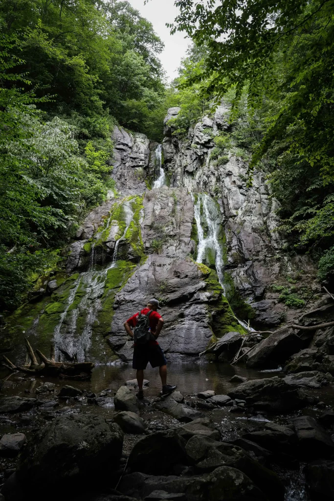 A hiker in a red shirt stands at the base of a rocky waterfall in Shenandoah National Park, surrounded by lush green forest. The cascading water flows gently over moss-covered rocks, creating a peaceful, hidden retreat deep in nature.