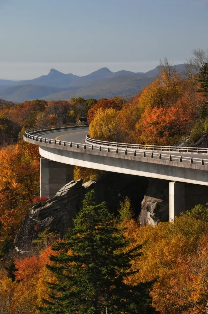 A curved bridge on the Blue Ridge Parkway cuts through a vibrant fall forest, with colorful autumn leaves in shades of orange, yellow, and red. The mountains in the background rise softly, creating a serene and majestic view, perfect for a peaceful road trip through nature.