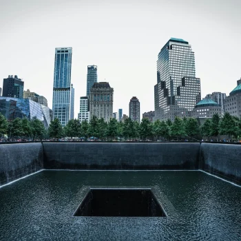 A view of the 9/11 Memorial in New York City, featuring the North Pool surrounded by trees and framed by the city's skyscrapers. The reflective pool, with its cascading water, serves as a somber tribute to the events of September 11, 2001.