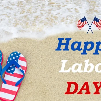 A pair of flip-flops with an American flag design lying on the sandy beach, with gentle ocean waves washing ashore. The image is accompanied by the text "Happy Labor Day!" in red, white, and blue, with two small American flags above the text.