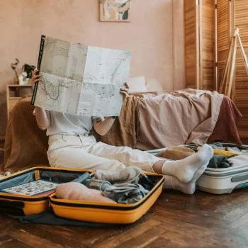 A person sitting on the floor surrounded by open suitcases, studying a large map in a cozy, softly lit bedroom. The scene captures the anticipation and preparation for an upcoming trip, with clothes, a hat, and travel essentials neatly packed.